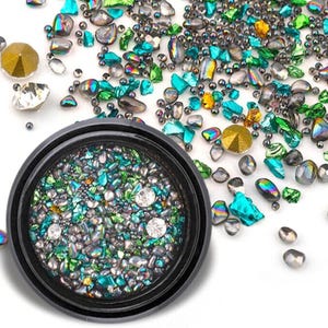 Puede incluir: Un recipiente negro lleno de pequeñas cuentas, cristales y gemas iridiscentes en tonos turquesa, verde, dorado y plateado. Alrededor del recipiente, hay cuentas y cristales sueltos adicionales, creando un efecto brillante. Ideal para nail art o proyectos de manualidades.