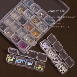 May include: A clear plastic jewelry box with multiple small compartments filled with various beads, gems, and pearls. The box is labeled "JEWELRY BOX" in white text. Some compartments are open, revealing the contents.