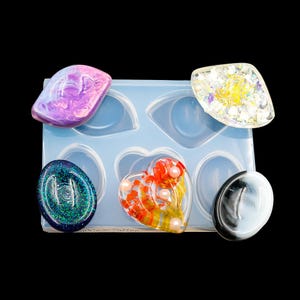 May include: A silicone mold with six cavities for making resin jewelry. The mold is blue and has a heart-shaped cavity, a round cavity, and an oval cavity. There are five resin pieces in the mold, including a purple swirl, a green glitter, a red and yellow flower, a white and black swirl, and a clear glitter.