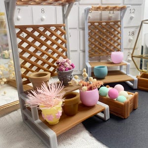 May include: Miniature garden display with two gray and brown shelves. Each shelf holds small pots with colorful plants and decorative spheres. The shelves have a lattice design and are set against a white background.