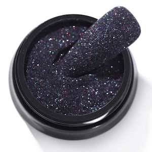 Puede incluir: Un frasco negro lleno de purpurina oscura. Se muestra una uña con purpurina encima del frasco. La purpurina tiene una base oscura con motas de colores iridiscentes, creando un efecto brillante. El frasco es redondo y negro.