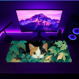 K&ouml;nnte beinhalten: Rechteckiges Mauspad mit einem Calico-Katzen-Design, das aus gr&uuml;nen Bl&auml;ttern hervorschaut. Die Katze hat orangefarbenes, wei&szlig;es und schwarzes Fell. Im Hintergrund befinden sich ein Computermonitor mit einer Berglandschaft und ein Computer-Tower.