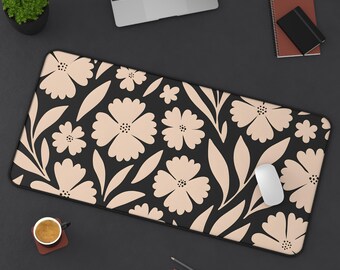 Puede incluir: Alfombrilla de escritorio negra con un diseño floral de flores y hojas beige. Un ratón de ordenador blanco descansa sobre la alfombrilla. La alfombrilla es rectangular y está diseñada para uso informático.