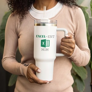 May include: A white insulated tumbler with a handle and a clear lid. The tumbler has the words "EXCEL-ENT MOM" printed in green. It includes a clear straw. The tumbler is held by a person.