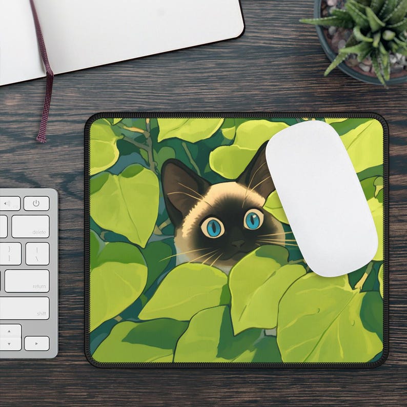 Puede incluir: Alfombrilla de rat&oacute;n rectangular con un gato siam&eacute;s mirando a trav&eacute;s de hojas verdes. El gato tiene ojos azules y una nariz negra. Un rat&oacute;n de ordenador blanco descansa sobre la alfombrilla, junto a un teclado y un cuaderno.