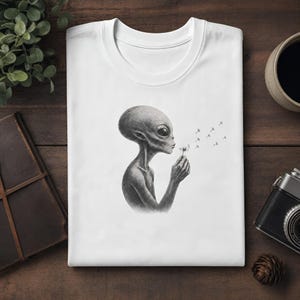 Könnte beinhalten: Ein weißes T-Shirt mit einer detaillierten schwarz-weißen Illustration eines Aliens, das auf eine Pusteblume bläst, wobei die Samen von der Blume wegfliegen. Die Grafik ist mittig auf der Brust platziert und zeigt den großen Kopf des Aliens.