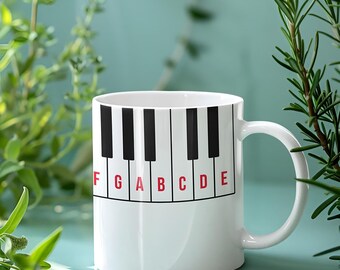 May include: White ceramic mug featuring a piano keyboard design. The black and white keys are labeled with red letters F, G, A, B, C, D, and E. The mug has a curved handle and is set against a light blue background with green foliage.