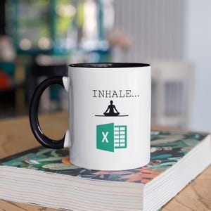 May include: White coffee mug with a black rim and handle. The mug has a green and black graphic of the Microsoft Excel logo with a person meditating above it. The text "INHALE..." is printed above the graphic.