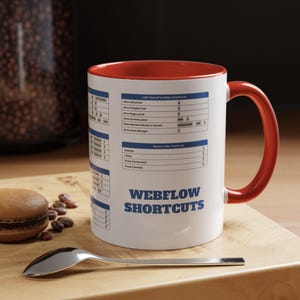 May include: White ceramic mug with a red rim and handle. The mug features a black and white graphic of Webflow keyboard shortcuts. The text "Webflow Shortcuts" is printed in bold blue letters on the mug.