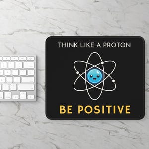 Op de afbeelding: Zwart rechthoekig muismat met de tekst "THINK LIKE A PROTON" en "BE POSITIVE" in wit en geel. De muismat heeft een cartoonatoom met een lachende blauwe kern en witte orbitale paden. Een wit toetsenbord is links zichtbaar.