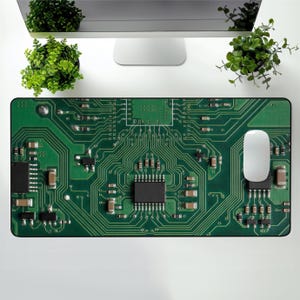 May include: A green rectangular mouse mat with a circuit board design. A white computer mouse sits on the right side. The mat's design includes electronic components and pathways, creating a tech-inspired aesthetic. The mat measures approximately 60 cm in length.