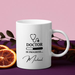 Könnte beinhalten: Weiße Keramiktasse mit schwarzem Stethoskop-Motiv, den Worten "DOCTOR IN PROGRESS..." und dem Namen "Michael". Die Tasse hat einen gebogenen Henkel und steht vor einem lila Hintergrund mit Orangenscheiben.