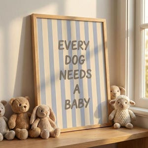 Op de afbeelding: Een ingelijste print met de tekst "EVERY DOG NEEDS A BABY" in grijze letters, tegen een blauw en crème gestreepte achtergrond. De lijst is van licht hout. Pluche speelgoed, waaronder een teddybeer, konijn en lam, is voor de print geplaatst.