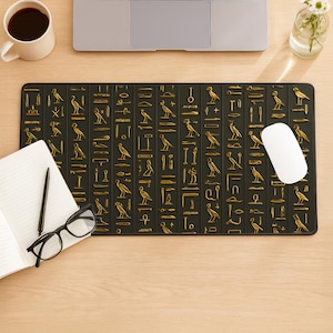 May include: Black rectangular mouse mat with gold hieroglyphic symbols. The design includes various bird and geometric shapes. A white mouse sits on the right side of the mat. The mat is on a wooden desk with a laptop, coffee, and a notebook.