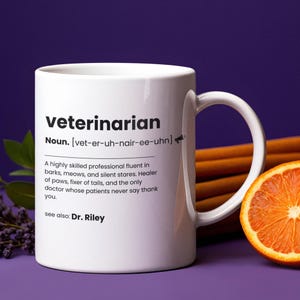 Peut inclure: Mug en céramique blanche avec la définition de "vétérinaire" imprimée en noir. La définition comprend la prononciation phonétique, une description du métier et "voir aussi : Dr. Riley". La tasse a une anse incurvée et est posée sur un fond violet.