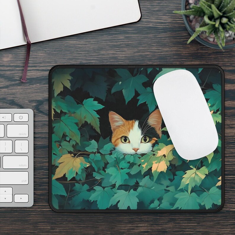 K&ouml;nnte beinhalten: Rechteckiges Mauspad mit einem Schildpatt-Katzenmotiv, das durch gr&uuml;ne und gelbe Bl&auml;tter blickt. Eine wei&szlig;e Computermaus liegt auf dem Pad. Das Mauspad hat einen schwarzen Rand.