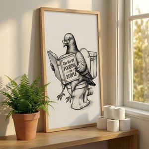 Pode incluir: Ilustração em preto e branco emoldurada de um pombo usando óculos e sentado em uma privada enquanto lê um livro intitulado "The Art of Pooping on People". A obra de arte é exibida em uma prateleira de madeira com um vaso de samambaia e rolos de papel higiênico.