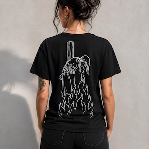 May include: A black short-sleeved t-shirt featuring a white line art graphic on the back. The design depicts a figure tied to a wooden post with ropes, positioned above stylised flames. The figure has long hair and appears to be smoking.