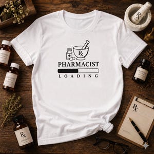 May include: A white short-sleeved t-shirt featuring a black graphic print of a mortar, pestle, and medicine bottle with the text PHARMACIST LOADING and a progress bar. The design includes the Rx symbol above the word PHARMACIST.