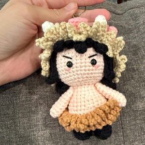 May include: A handmade crochet doll keychain featuring a character with black hair, pink cheeks, and a tan and pink headpiece. The doll has a light pink body, a tan skirt, and black shoes. The keychain is attached to a silver metal ring.