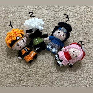 May include: Four handmade crochet dolls, each depicting a different character. The dolls feature vibrant colors: orange, black, white, green, blue, and pink. The dolls are arranged on a neutral-colored surface. The dolls are approximately 4 inches tall.