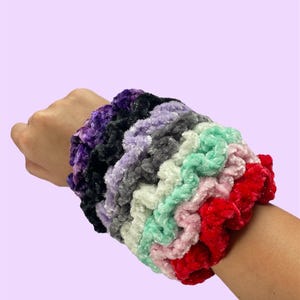 May include: A stack of colorful, fuzzy hair scrunchies in various shades of purple, black, gray, white, pink, green, and red.