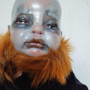 May include: A close-up of a doll's face with a pale, gray complexion and red accents. The doll has a bald head, dark eyes, and a reddish-brown fur collar. The doll is wearing a black garment.