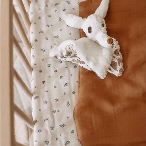 Peut inclure: Un sac de couchage brun avec un jouet en peluche de chauve-souris blanc à l'intérieur. Le sac de couchage est sur un drap de lit blanc avec un petit motif floral.