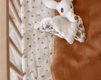 May include: A brown sleep sack with a white plush toy bat inside. The sleep sack is on a white crib sheet with a small floral pattern.