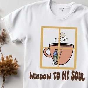 Könnte beinhalten: Weißes T-Shirt mit einem Grafikdruck einer Kaffeetasse mit aufsteigendem Dampf. Die Tasse befindet sich in einem Fensterrahmen, und eine Person klettert auf einer Leiter in der Tasse. Der Text "Window to my soul" ist unter dem Grafikdruck aufgedruckt.