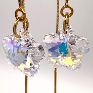 Puede incluir: Un par de pendientes de cadena dorados con colgantes de cristal transparente facetado en forma de flor. Los pendientes cuelgan de una rama de madera sobre un fondo suave y neutro. Los cristales reflejan múltiples colores.