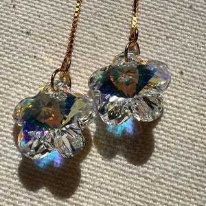 May include: A pair of clear, faceted crystal flower earrings with gold-colored chains. The earrings are shaped like five-petal flowers and reflect a rainbow of colors. The earrings are hanging against a neutral, textured background.