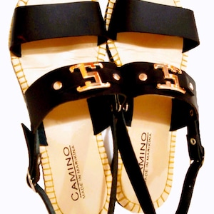 May include: A pair of black sandals with two wide straps across the top, featuring a gold-toned metal emblem on the front strap. The sandals have a light-colored footbed with the text CAMINO MADE IN MARIKINA and a slingback strap with a buckle.