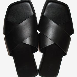 May include: A pair of black slide sandals featuring a square toe design and two wide, overlapping straps that cross over the top of the foot. The footbed is flat and black, with a smooth finish and a visible brand logo embossed on the heel area.