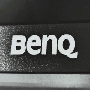 Könnte beinhalten: Nahaufnahme einer schwarzen Oberfläche mit dem weißen Wort "BenQ", gedruckt in einer fetten, serifenlosen Schriftart. Die Buchstaben sind gleichmäßig angeordnet und vor dem dunklen Hintergrund gut sichtbar. Das Bild ist gut beleuchtet.