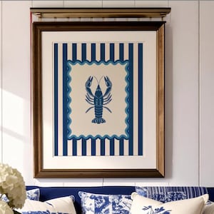 Op de afbeelding: Een ingelijste print met een blauw kreeftontwerp op een crèmekleurige achtergrond, tegen een gestreept blauw-wit patroon. Het kunstwerk wordt weergegeven boven een blauwe bank met decoratieve kussens. Twee messing wandkandelaars flankeren het kunstwerk.