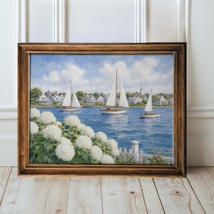 May include: A framed painting shows a tranquil coastal scene. Three sailboats sail on blue water, with white houses and a church in the background under a cloudy sky. White flowers and a white picket fence are in the foreground.