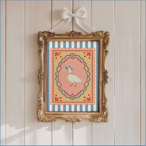 May include: An ornate gold framed artwork. The artwork features a white goose wearing a blue hat and red bow. The background has blue and white stripes, with a pink oval frame. The artwork is suspended by a white ribbon bow.