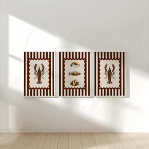May include: Three framed art prints featuring marine life. Two prints display a brown lobster illustration, while the center print showcases three colorful fish. Each print has a cream-colored background with a brown scalloped border and a striped frame.