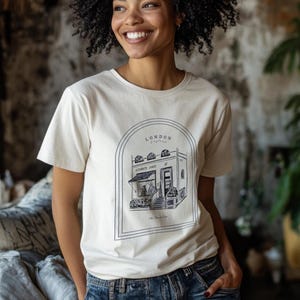 Può includere: Una maglietta bianca con un'illustrazione in bianco e nero di un edificio con il testo "LONDON England" e "Elizabeth James" sopra la porta. Il testo "The Flower Shop" è sotto l'edificio.