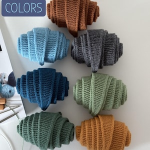 Pode incluir: Uma coleção de itens em forma de croissant, feitos à mão, em crochê, em várias cores, incluindo marrom, azul, cinza, verde e laranja. A imagem inclui o texto "NEW COLORS" em uma caixa azul. Estes itens parecem ser decorativos ou de novidade.