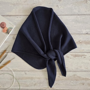 Può includere: Uno scialle lavorato a maglia blu navy, annodato sul davanti. Lo scialle è ripiegato, creando una forma triangolare. Ferri da maglia e un libro con campioni di filato sono visibili sullo sfondo. Lo scialle è realizzato in materiale morbido e strutturato.