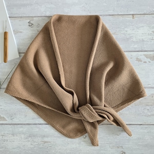 May include: A knitted shawl in a tan hue, knotted at the front. The shawl is arranged to display its texture and shape. The background is a light-coloured wooden surface. A crochet hook is visible in the upper left corner.