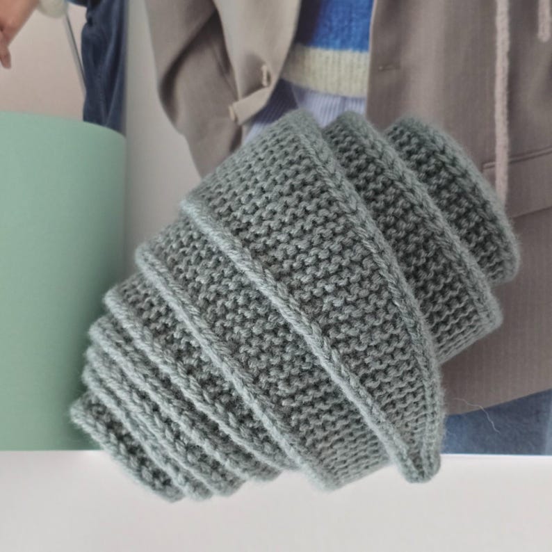 Op de afbeelding: Een grijze gebreide sjaal met een geribbelde textuur. De sjaal is spiraalvormig gevouwen.