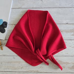 May include: A vibrant red knitted shawl, folded and tied at the front. The shawl is made of a soft, textured material and is displayed on a light-colored wooden surface. The shawl's design is triangular, with long ties.