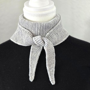 Op de afbeelding: Een grijze gebreide nekwarmer met een strik aan de voorkant. De nekwarmer zit op een witte mannequin.