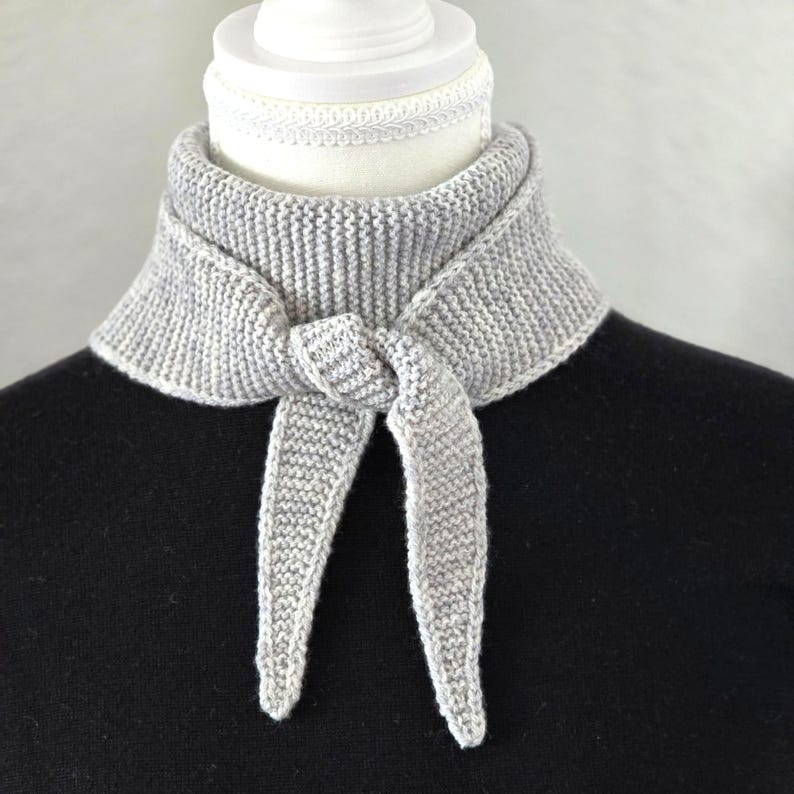 Op de afbeelding: Een grijze gebreide nekwarmer met een strik aan de voorkant. De nekwarmer zit op een witte mannequin.