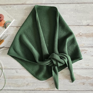 May include: A forest green knitted shawl, knotted at the front. The shawl is made of a textured fabric and is folded on a light wood surface. Knitting needles and yarn are visible in the background.