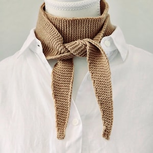 May include: A tan knitted scarf tied around a white shirt collar. The scarf is made of a soft, textured yarn and has a simple, elegant design.
