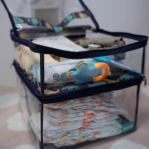 May include: Clear plastic storage bin with a navy blue zipper and a dinosaur print. The bin is filled with baby items, including nappies, wipes, and a toy.